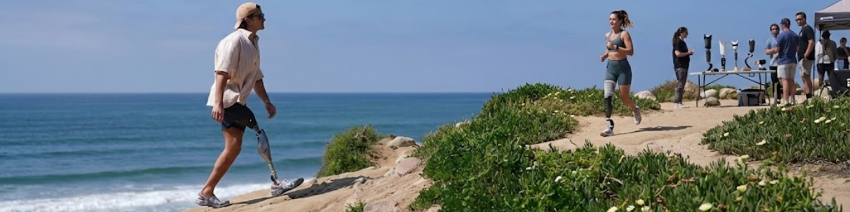 Mężczyzna i kobieta z nowoczesnymi protezami nóg aktywnie poruszający się na nadmorskim klifie. Mężczyzna schodzi po zboczu, kobieta biegnie ścieżką; w tle stanowisko z ekspozycją różnych modeli protez kończyn dolnych.