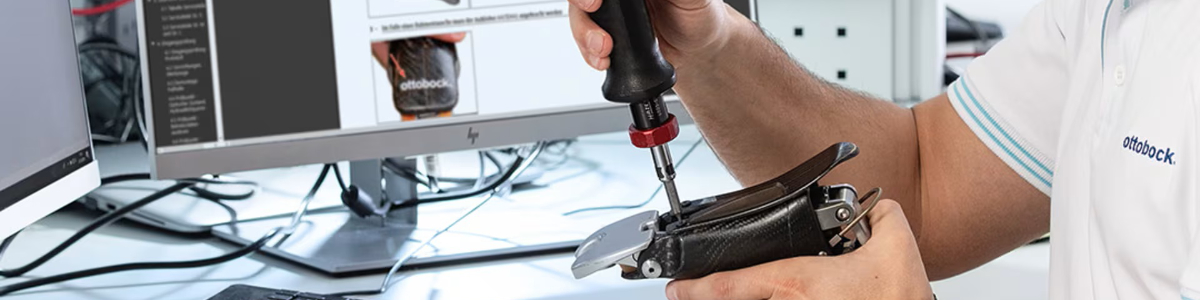 Technik ortopeda wykonuje przegląd techniczny mechatronicznej stopy protetycznej marki Ottobock przy użyciu klucza dynamometrycznego, w tle monitor z instrukcją serwisową.